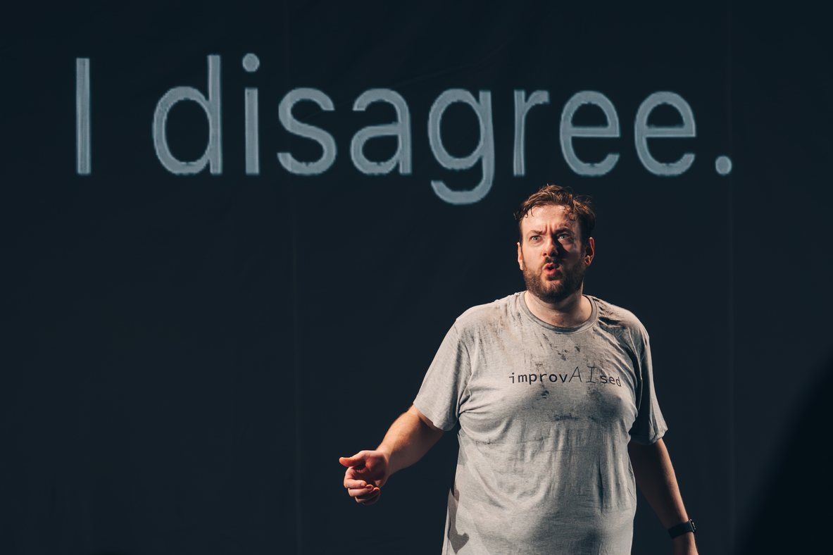 Tom Gilder performing beneath projected text reading I disagree during ImprovAIsed